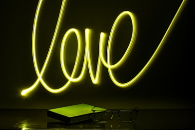 Neon light spelling out the word 'love' in bright yellow against a dark background. Below it, a closed book and a pair of eyeglasses are placed on a reflective surface, casting subtle reflections.