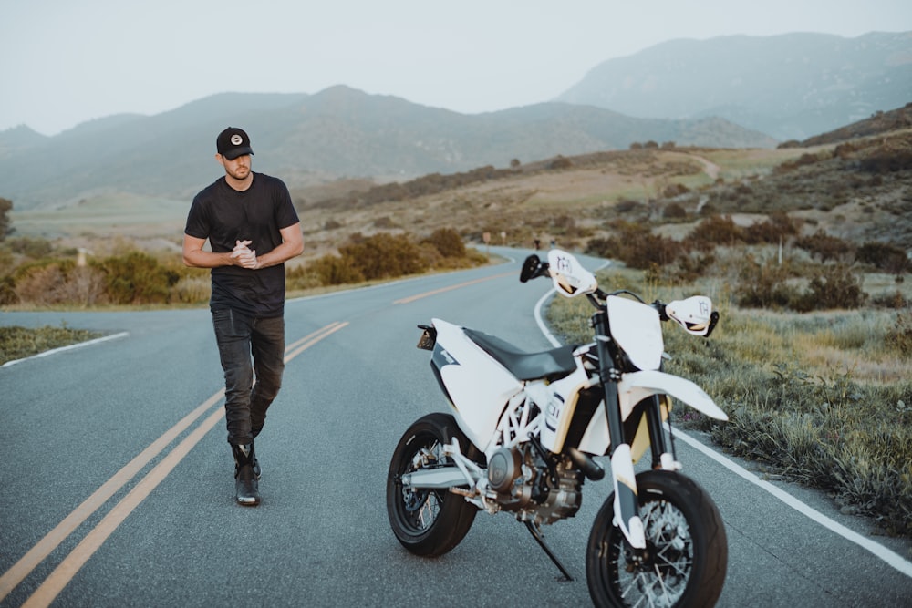 man in black sweater and blue denim jeans standing beside white and black motorcycle during daytime