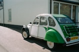 A vintage green and white car is parked on the asphalt near a building with white walls and large windows. The car has a classic rear design, featuring a distinctive shape and a European license plate.