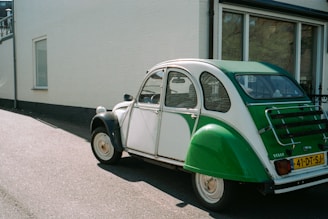 A vintage green and white car is parked on the asphalt near a building with white walls and large windows. The car has a classic rear design, featuring a distinctive shape and a European license plate.