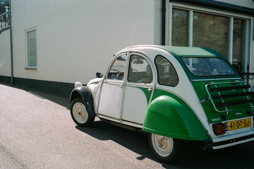 A vintage green and white car is parked on the asphalt near a building with white walls and large windows. The car has a classic rear design, featuring a distinctive shape and a European license plate.