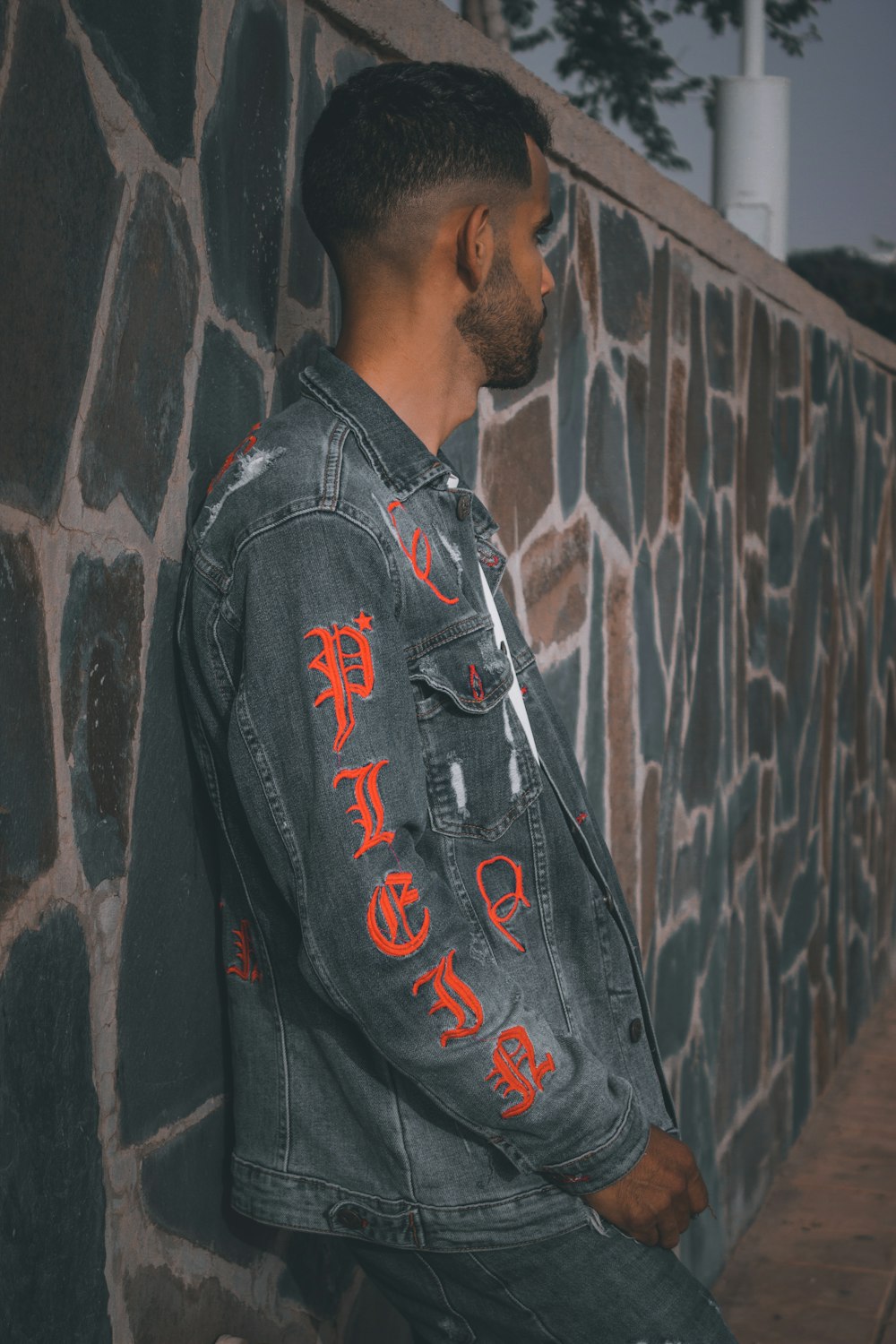 a man leaning against a stone wall wearing a denim jacket