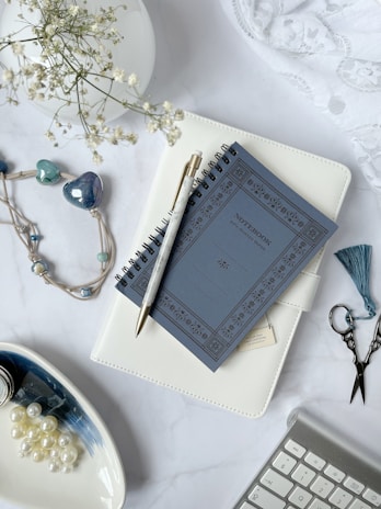 A stylish flat lay features a spiral notebook with a dark blue cover placed on top of a white planner. A gold and white pen rests on the notebook. Nearby is a beaded necklace with heart-shaped accents, a small pair of ornate scissors, and a tassel. White flowers in a vase add a decorative touch. Pearls are displayed in a dish alongside a black ink bottle. A keyboard appears at the edge of the composition.