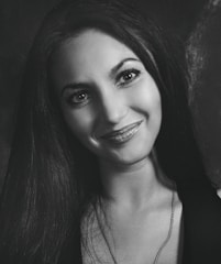 A black and white portrait of a smiling woman with long dark hair and bright eyes. She is wearing a necklace and her expression appears friendly and inviting.
