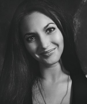 A black and white portrait of a smiling woman with long dark hair and bright eyes. She is wearing a necklace and her expression appears friendly and inviting.