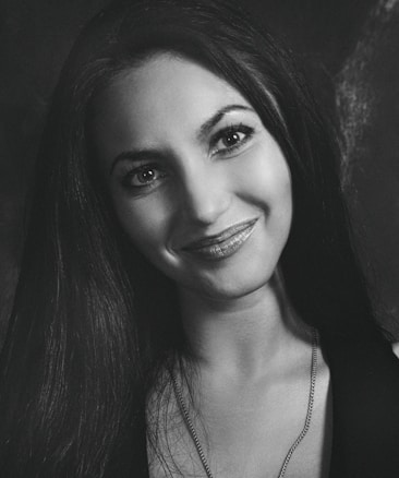 A black and white portrait of a smiling woman with long dark hair and bright eyes. She is wearing a necklace and her expression appears friendly and inviting.