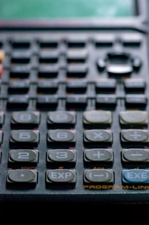 Close-up view of a calculator with numerous buttons, including numbers, arithmetic operations, and special functions like EXP and EXE. The keys are dark in color, with white labels, and the surface has a slightly worn appearance.