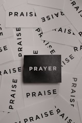 A collection of white cards spread out with the word 'PRAISE' printed in bold black letters. In the center, there is a single black card with the word 'PRAYER' in white letters.
