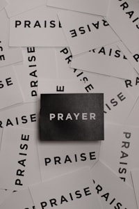 A collection of white cards spread out with the word 'PRAISE' printed in bold black letters. In the center, there is a single black card with the word 'PRAYER' in white letters.