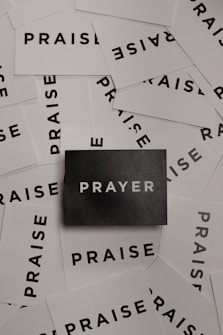 A collection of white cards spread out with the word 'PRAISE' printed in bold black letters. In the center, there is a single black card with the word 'PRAYER' in white letters.