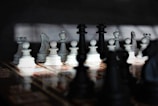A chessboard with an arranged set of white and black chess pieces is situated on a patterned surface. The background is shadowed, with light casting highlights on some pieces, creating a dramatic effect.