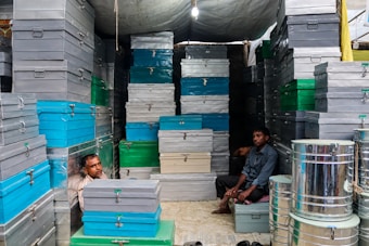 A small shop or storage area with stacks of metal boxes in various sizes and colors, including gray, blue, and green. Two men are seated amongst the boxes, one on the left and the other on the right. The lighting is artificial and slightly dim.