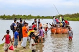 A group of people are gathered near a riverbank, some standing on the grassy shore while others are getting off a small boat. The main focus is on the orange boat filled with people, which is docked at the river's edge. Some individuals are wading through knee-deep water to reach the boat. The background features lush green trees and a cloudy sky.