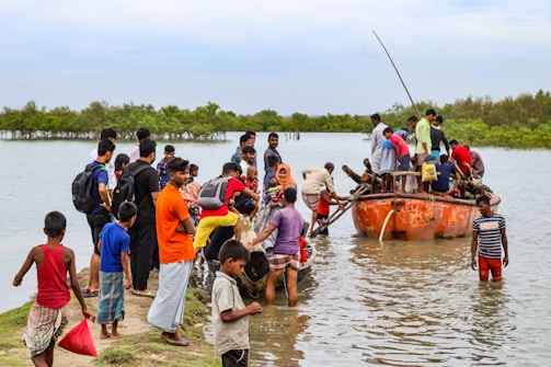 A group of people are gathered near a riverbank, some standing on the grassy shore while others are getting off a small boat. The main focus is on the orange boat filled with people, which is docked at the river's edge. Some individuals are wading through knee-deep water to reach the boat. The background features lush green trees and a cloudy sky.