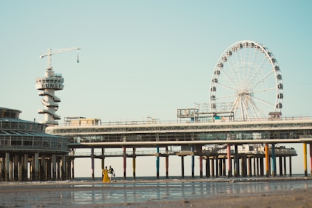 A beachside scene with a tall bungee tower labeled 'BUNGY' to the left, and a large Ferris wheel to the right. Both structures are on a long pier stretching across the image with colorful pillars. The sandy beach is in the foreground, with two people walking near the water's edge.