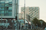 Two modern apartment buildings with a complex of balconies stand adjacent to a busy urban street. Various traffic lights and signs are visible, along with a cyclist passing by. A tram line with overhead wires can be seen, indicating a public transportation area, and there are several trees lining the street.