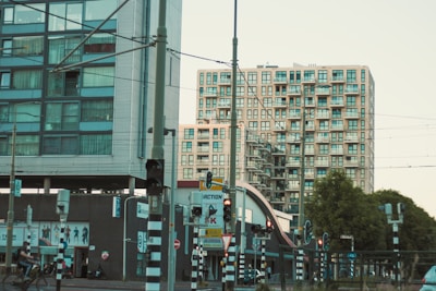 Two modern apartment buildings with a complex of balconies stand adjacent to a busy urban street. Various traffic lights and signs are visible, along with a cyclist passing by. A tram line with overhead wires can be seen, indicating a public transportation area, and there are several trees lining the street.