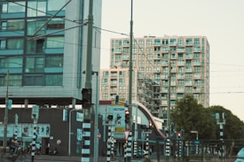 Two modern apartment buildings with a complex of balconies stand adjacent to a busy urban street. Various traffic lights and signs are visible, along with a cyclist passing by. A tram line with overhead wires can be seen, indicating a public transportation area, and there are several trees lining the street.