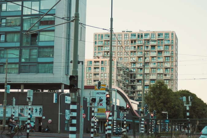 Two modern apartment buildings with a complex of balconies stand adjacent to a busy urban street. Various traffic lights and signs are visible, along with a cyclist passing by. A tram line with overhead wires can be seen, indicating a public transportation area, and there are several trees lining the street.