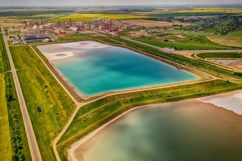 An aerial view of an industrial landscape featuring large water bodies with vibrant turquoise and brown hues. The surrounding area includes green fields and a complex of red-roofed industrial buildings. Roads and pathways can be seen bordering the water bodies and fields.