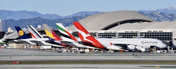 A lineup of large passenger airplanes with distinctive airline branding are parked at an airport terminal with a mountainous backdrop. The scene includes several planes from different international airlines, positioned adjacent to one another, showcasing their colorful tail designs.