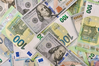 A collection of various currency notes, including US dollars and euros, spread out and overlapping each other. The notes display intricate designs with portraits and architectural images.