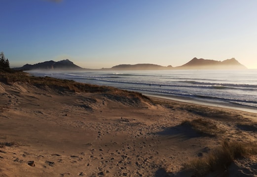 A serene beach scene at sunrise or sunset, featuring light golden sand, gentle waves, and distant mountains under a clear blue sky. Footprints are visible on the sand, leading toward the water, and a few small figures are surfing in the ocean.