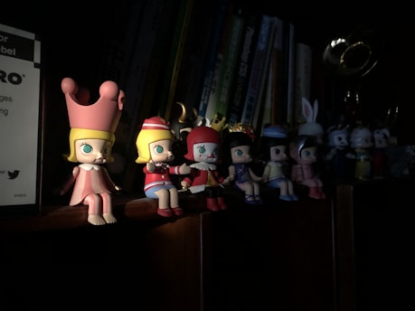 A set of small, colorful figurines depicting cartoon-like characters with exaggerated features are arranged on a wooden shelf. Each character wears distinct outfits, such as crowns and hats. The shelf is part of a bookcase with several books visible in the background. The lighting is dim, creating a dramatic shadow effect on the figurines.