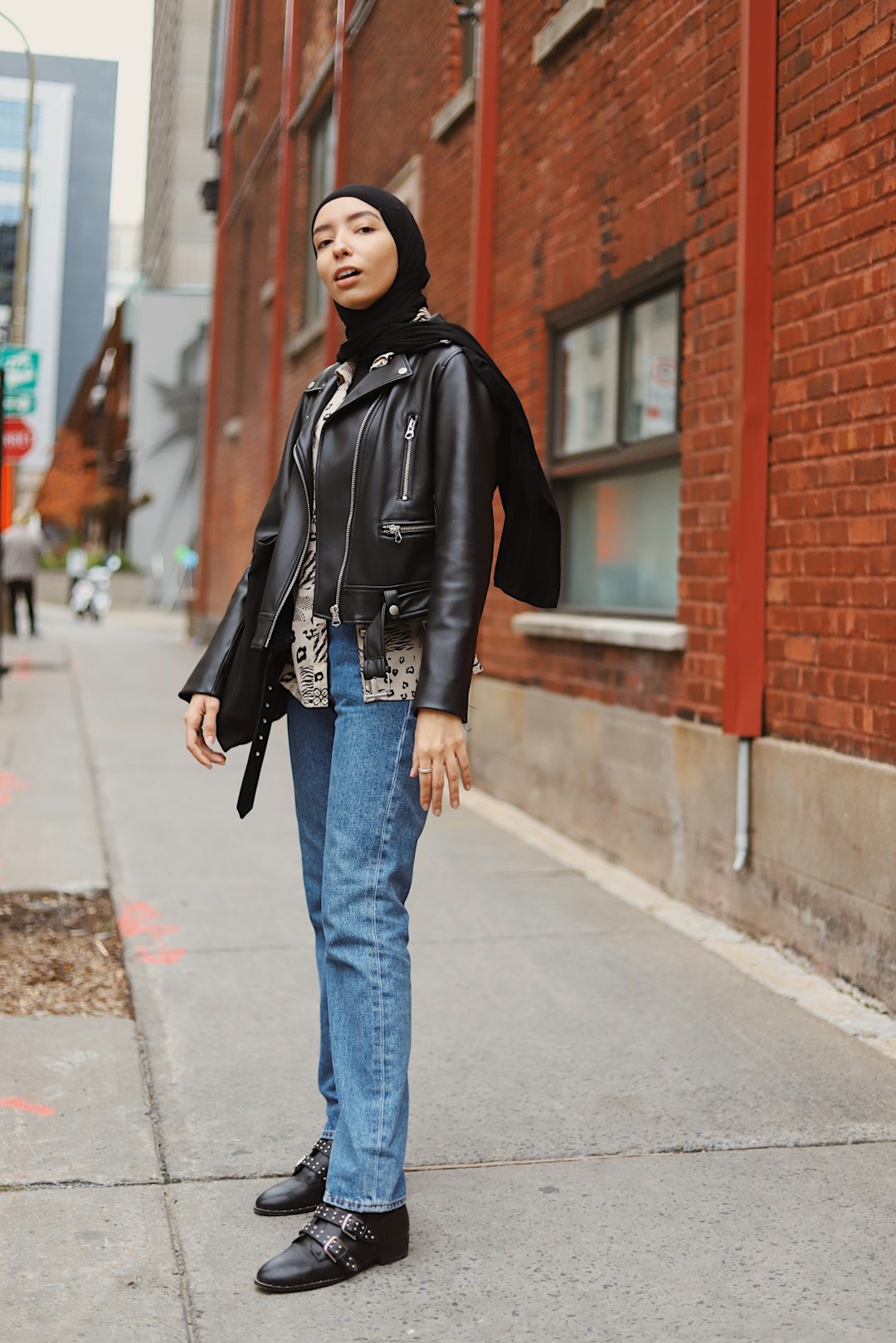 woman in black leather jacket and blue denim jeans standing on sidewalk during daytime