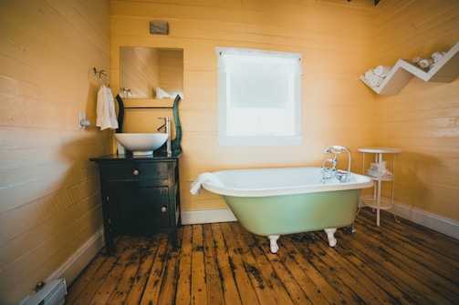A rustic bathroom features wooden plank flooring and light yellow walls. There is a vintage-style green freestanding bathtub with chrome fixtures, positioned near a window covered by a white curtain. Adjacent to the tub, a white round table holds a plant and towels. A black vanity cabinet with a white vessel sink and a large rectangular mirror is placed on the left side. Towels hang on a hook next to the sink.
