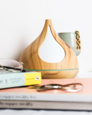 A wooden aromatherapy diffuser with a teardrop design is placed on a surface next to a stack of books. A pair of sunglasses rests on the books, and a ceramic mug with a gold chain lies behind the diffuser.