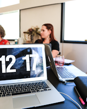 A close-up of a workspace featuring an open laptop displaying a large digital clock reading 12:17. In the background, two women are sitting at a table, engaging in conversation or a meeting. The setting appears to be an office or a study space with natural light streaming in from a window. Additional items, including another laptop, a glass, and a notebook with a pen, are visible on the table.