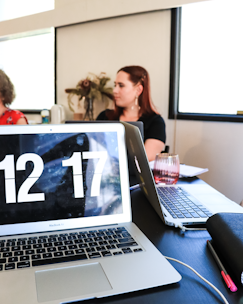 A close-up of a workspace featuring an open laptop displaying a large digital clock reading 12:17. In the background, two women are sitting at a table, engaging in conversation or a meeting. The setting appears to be an office or a study space with natural light streaming in from a window. Additional items, including another laptop, a glass, and a notebook with a pen, are visible on the table.