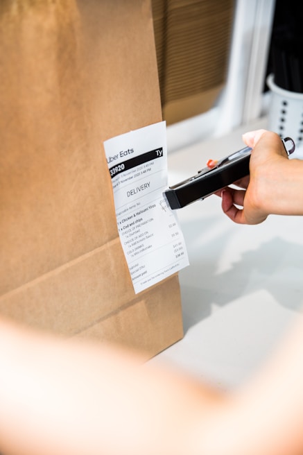 A hand is holding a scanner over a delivery receipt attached to a brown paper bag. The setting appears to be a workspace or a kitchen counter.