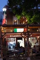 An outdoor dining area at a pizzeria with people sitting at tables in the evening. The restaurant features a wooden structure with string lights and an Italian flag prominently displayed. There is a lot of greenery surrounding the area, and the interior of the pizzeria is visible with staff and customers inside.