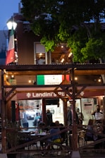 An outdoor dining area at a pizzeria with people sitting at tables in the evening. The restaurant features a wooden structure with string lights and an Italian flag prominently displayed. There is a lot of greenery surrounding the area, and the interior of the pizzeria is visible with staff and customers inside.
