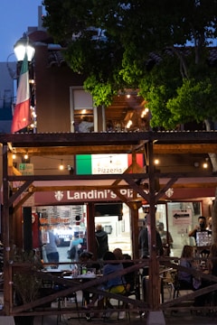 An outdoor dining area at a pizzeria with people sitting at tables in the evening. The restaurant features a wooden structure with string lights and an Italian flag prominently displayed. There is a lot of greenery surrounding the area, and the interior of the pizzeria is visible with staff and customers inside.