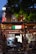An outdoor dining area at a pizzeria with people sitting at tables in the evening. The restaurant features a wooden structure with string lights and an Italian flag prominently displayed. There is a lot of greenery surrounding the area, and the interior of the pizzeria is visible with staff and customers inside.