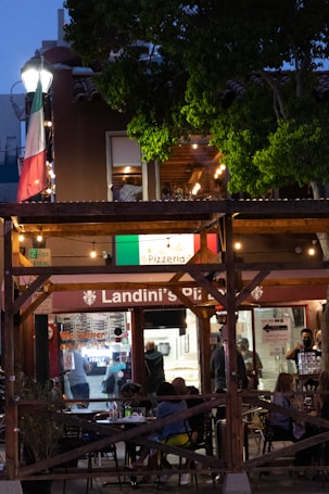 An outdoor dining area at a pizzeria with people sitting at tables in the evening. The restaurant features a wooden structure with string lights and an Italian flag prominently displayed. There is a lot of greenery surrounding the area, and the interior of the pizzeria is visible with staff and customers inside.