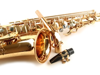 A close-up view of a shiny gold saxophone with intricate details and silver keys. The image also features a black mouthpiece with a metal ligature placed in front of the saxophone. The background is plain and white, highlighting the instrument's gleaming surface.