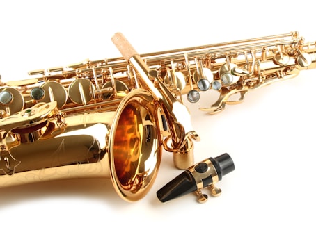 A close-up view of a shiny gold saxophone with intricate details and silver keys. The image also features a black mouthpiece with a metal ligature placed in front of the saxophone. The background is plain and white, highlighting the instrument's gleaming surface.