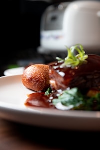 A close-up of a gourmet dish featuring a piece of glazed meat garnished with fresh herbs, served with a crispy, golden-brown croquette, and accompanied by green vegetables. The plate is set on a wooden surface with a blurred background, possibly a kitchen appliance.