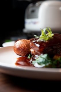 A close-up of a gourmet dish featuring a piece of glazed meat garnished with fresh herbs, served with a crispy, golden-brown croquette, and accompanied by green vegetables. The plate is set on a wooden surface with a blurred background, possibly a kitchen appliance.