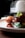 A close-up of a gourmet dish featuring a piece of glazed meat garnished with fresh herbs, served with a crispy, golden-brown croquette, and accompanied by green vegetables. The plate is set on a wooden surface with a blurred background, possibly a kitchen appliance.