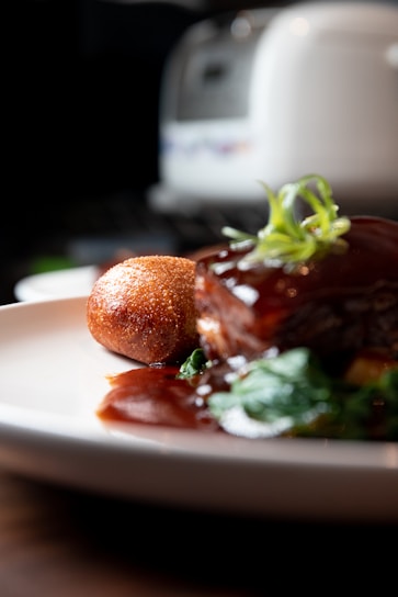 A close-up of a gourmet dish featuring a piece of glazed meat garnished with fresh herbs, served with a crispy, golden-brown croquette, and accompanied by green vegetables. The plate is set on a wooden surface with a blurred background, possibly a kitchen appliance.
