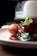 A close-up of a gourmet dish featuring a piece of glazed meat garnished with fresh herbs, served with a crispy, golden-brown croquette, and accompanied by green vegetables. The plate is set on a wooden surface with a blurred background, possibly a kitchen appliance.