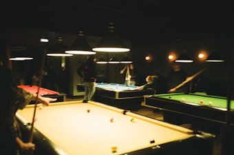 Several people are playing pool in a dimly lit room with multiple pool tables. The lighting is warm, coming from hanging overhead lamps. Various colors of pool balls are scattered across the tables, and cues are visible in the hands of some players.