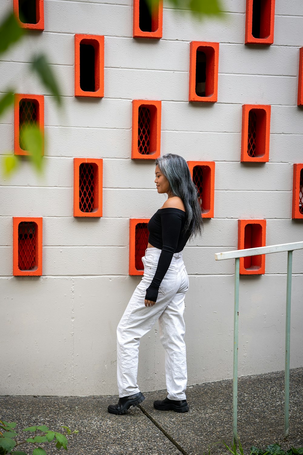 Mujer con camisa negra de manga larga y pantalones blancos de pie junto a la pared roja y blanca