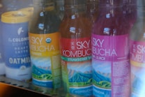 Several bottles of Sky Kombucha in various flavors are lined up in a refrigerator. The bottles have colorful labels with images of mountains and are placed next to a tall can of La Colombe Coffee Draft Latte made with oat milk.
