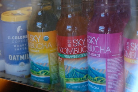 Several bottles of Sky Kombucha in various flavors are lined up in a refrigerator. The bottles have colorful labels with images of mountains and are placed next to a tall can of La Colombe Coffee Draft Latte made with oat milk.